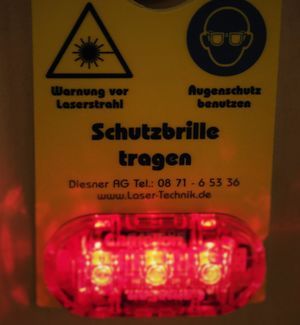 A warning light at the entrance to the laser laboratory is shown.  The sign laser warns against emitting visible and invisible laser irradiation. Always wear safety glasses.