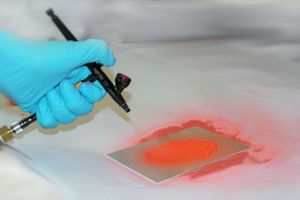 The illustration shows the application of a red liquid on a metal sheet with an airbrush gun.