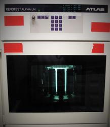 Die Abbildung zeigt den Xenotester Beta+. Die Testkammer ist in Betrieb und man sieht die Proben auf einen Probenkarusell um die hell erleuchtete Strahlungsquelle im Mittelpunkt fahren.