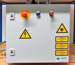 The figure shows the control box of the CO2 laser workstation. There is an emergency stop switch, other switches and many warning signs to be seen. 